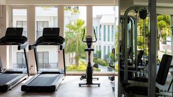 Un gimnasio con cintas de correr, una bicicleta estática y máquinas de pesas. Cuenta con ventanas amplias que ofrecen vistas a un edificio y zonas verdes.