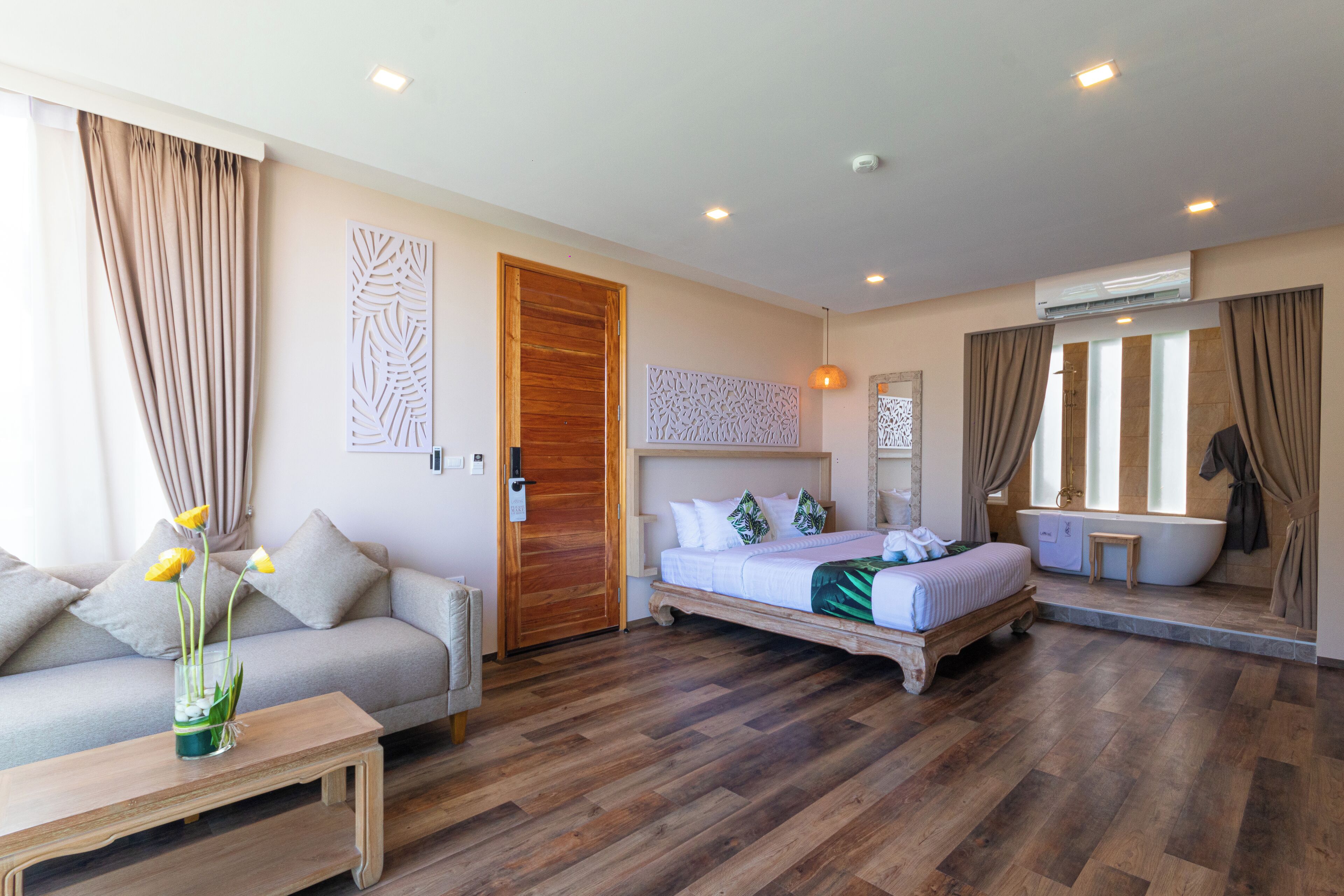 A hotel room with a wooden floor, a bed with white bedding, a sofa, a small table with a vase of flowers, and a bathroom with a bathtub and shower.