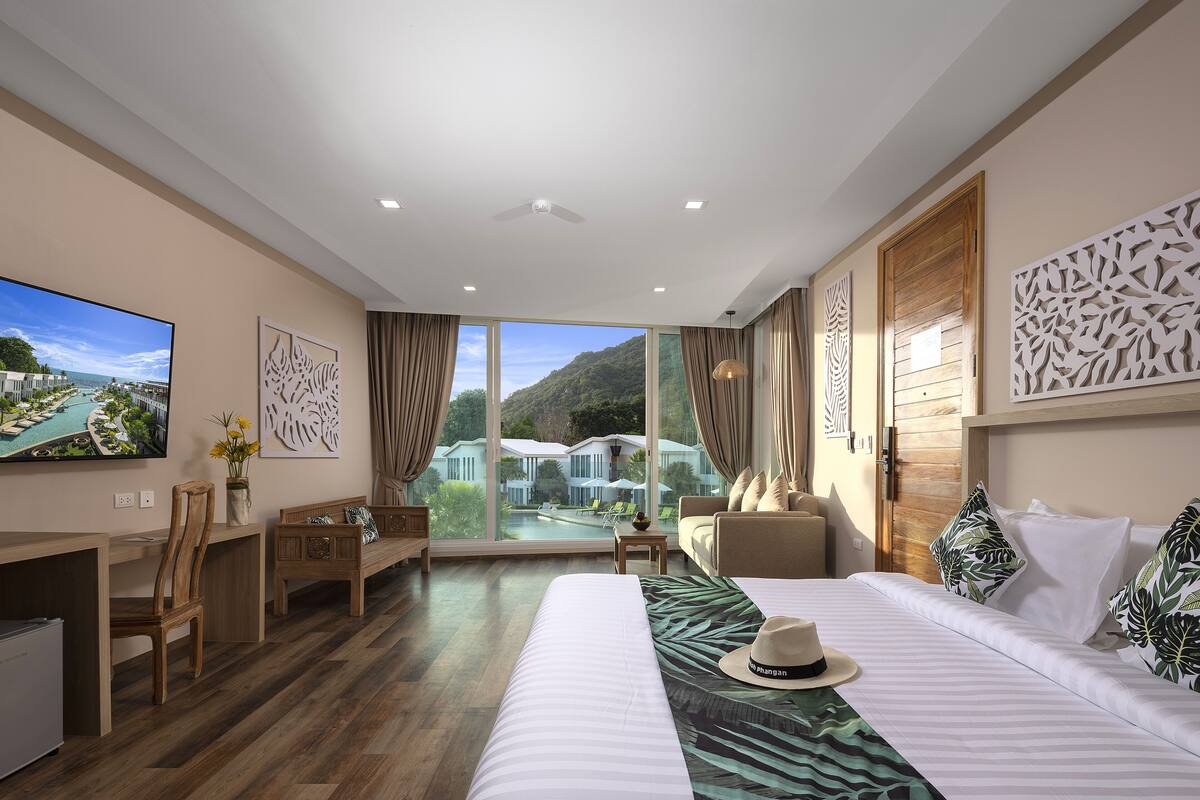 A hotel room with a large bed, a desk, a chair, a TV, and a view of a pool and mountains.