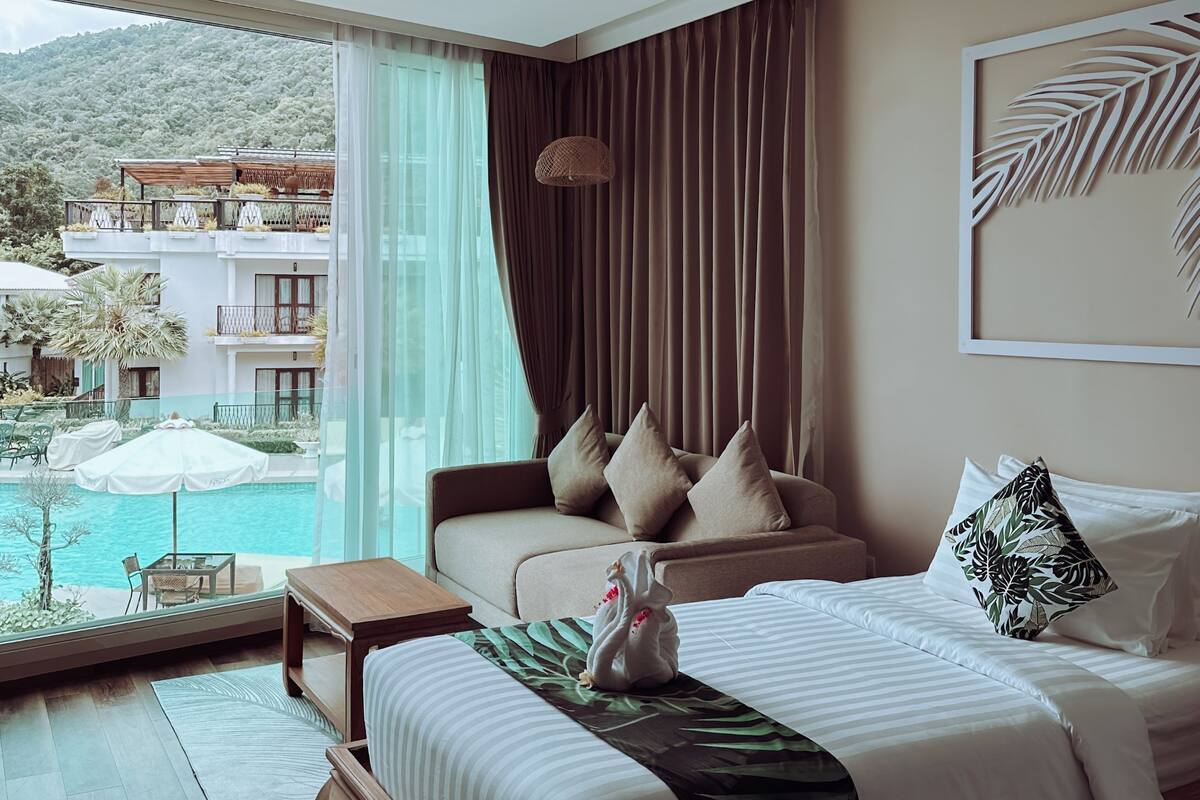 A hotel room with a bed, a view of a pool and building, and a ceiling light.