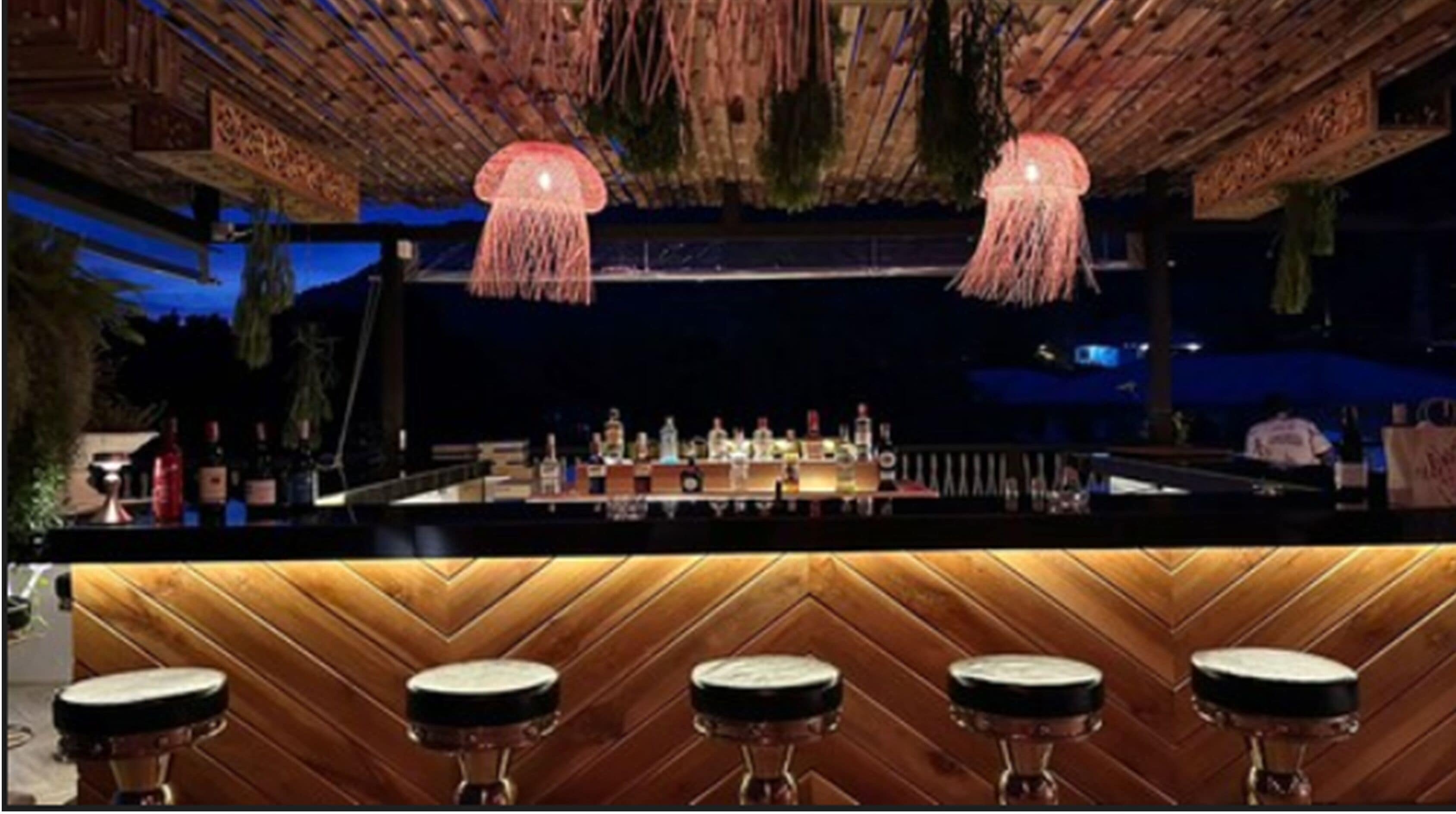 A bar area with a wooden bar counter, hanging pendant lights, and stools.