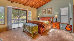 Ein Schlafzimmer mit Holzbalkendecke, einem Bett mit bunter Decke, einem Schminktisch, einer Gitarre und einem Fenster mit Jalousien.