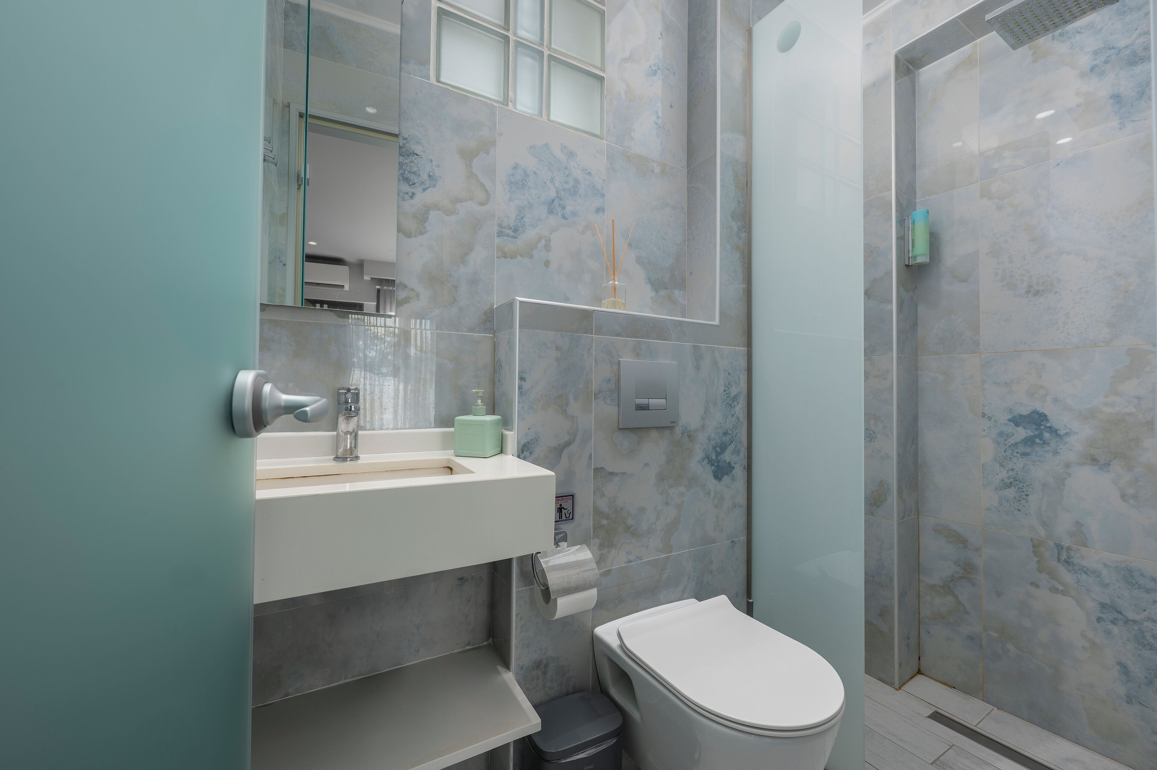 Ein modernes Badezimmer mit Duschkabine aus Glas, einem weißen Waschbecken mit grünem Seifenspender und einer Toilette mit Papiertuchspender.