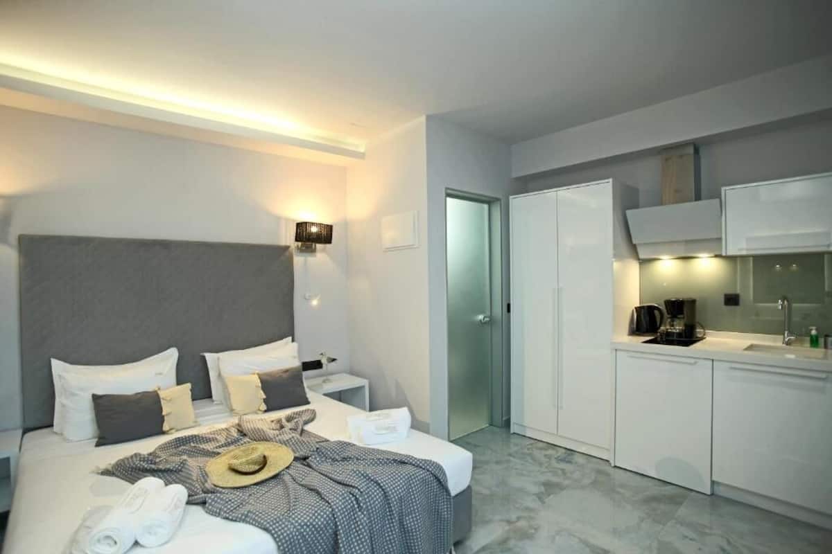 A modern hotel room with a large bed, a small bedside table, a desk, a kitchenette, and a wardrobe.