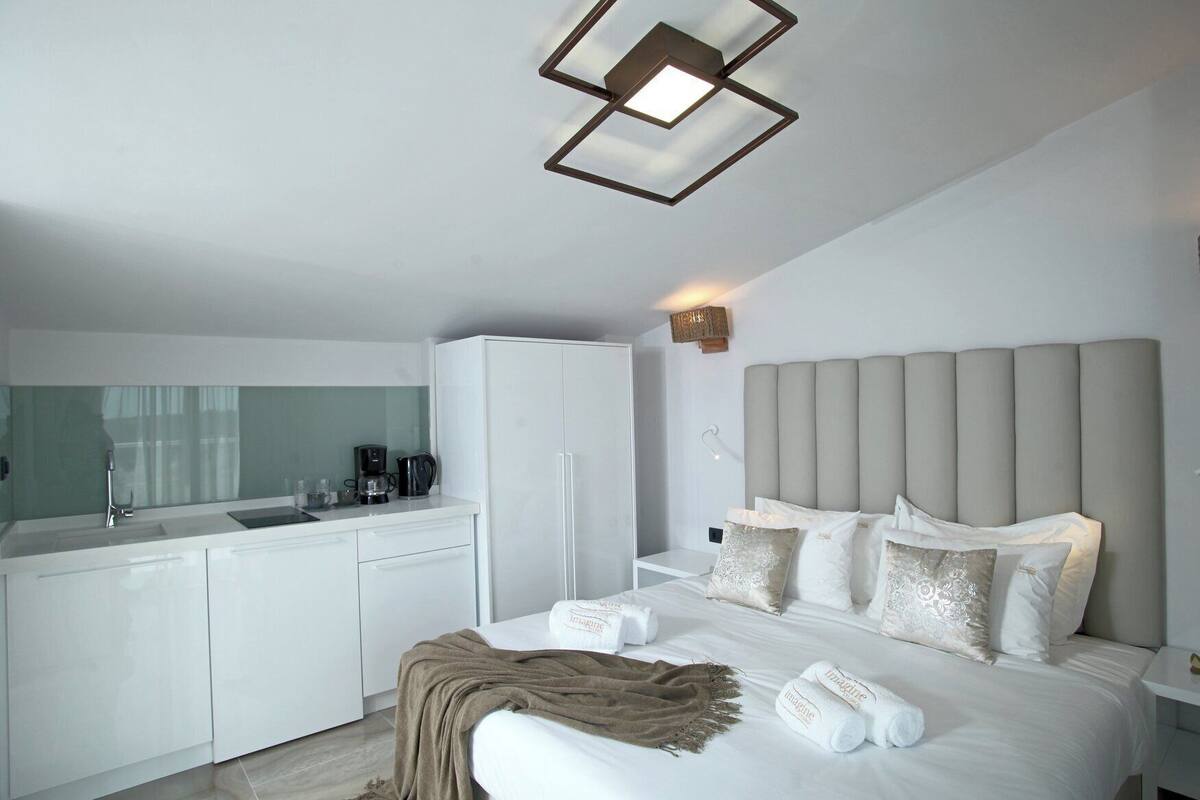 A modern hotel room with a white bed, a beige headboard, and a kitchen area with a white refrigerator and a coffee maker.