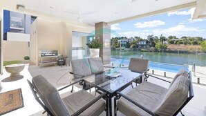 Un moderno comedor al aire libre con una mesa y sillas de vidrio, con vista a una piscina y un paisaje con casas y follaje.