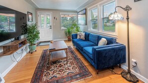 Un salon avec un canapé bleu, une table basse en bois et un tapis à motifs. On y trouve deux plantes en pot, un téléviseur posé sur un meuble en bois et une lampe sur pied.