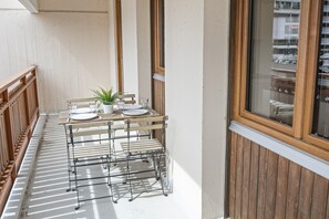 A balcony with a table set for two, a potted plant, and a window with blinds. - Appartement central et confortable (Crans (VS))