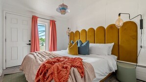 A bedroom with a bed, a bedside lamp, a window with curtains, and a view of the outdoors. - The Mango  Vibrant Oasis Sleeps 4  Downtown Fun (Drumheller)