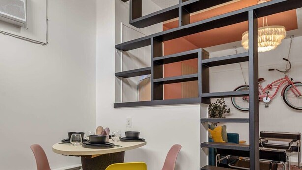 A dining area with a round table, two chairs, and a modern bookshelf with a bicycle. - The Mango  Vibrant Oasis Sleeps 4  Downtown Fun (Drumheller)