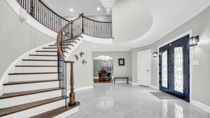 A spacious foyer with a curved staircase, a dining area, and a dark blue door with decorative glass panels. - The Wexford French Chateau w/ Pool (Athens)