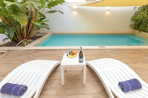 Un espace aménagé au bord de la piscine, avec des chaises longues, une table sur laquelle se trouvent une bouteille et des fruits, ainsi que des plantes.