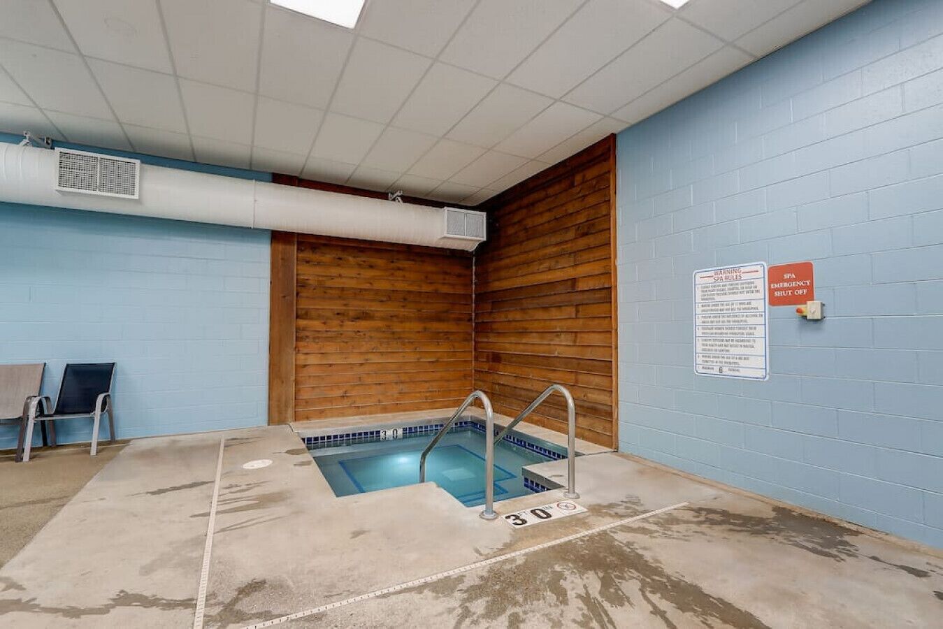 A small indoor pool with a wooden wall and a blue tiled floor. There are two chairs and a sign on the wall.