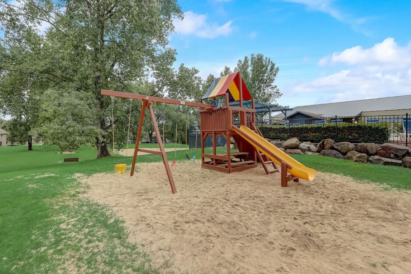 Une aire de jeux avec un toboggan et des balançoires, entourée de sable et d’herbe, avec des arbres et un bâtiment en arrière-plan.