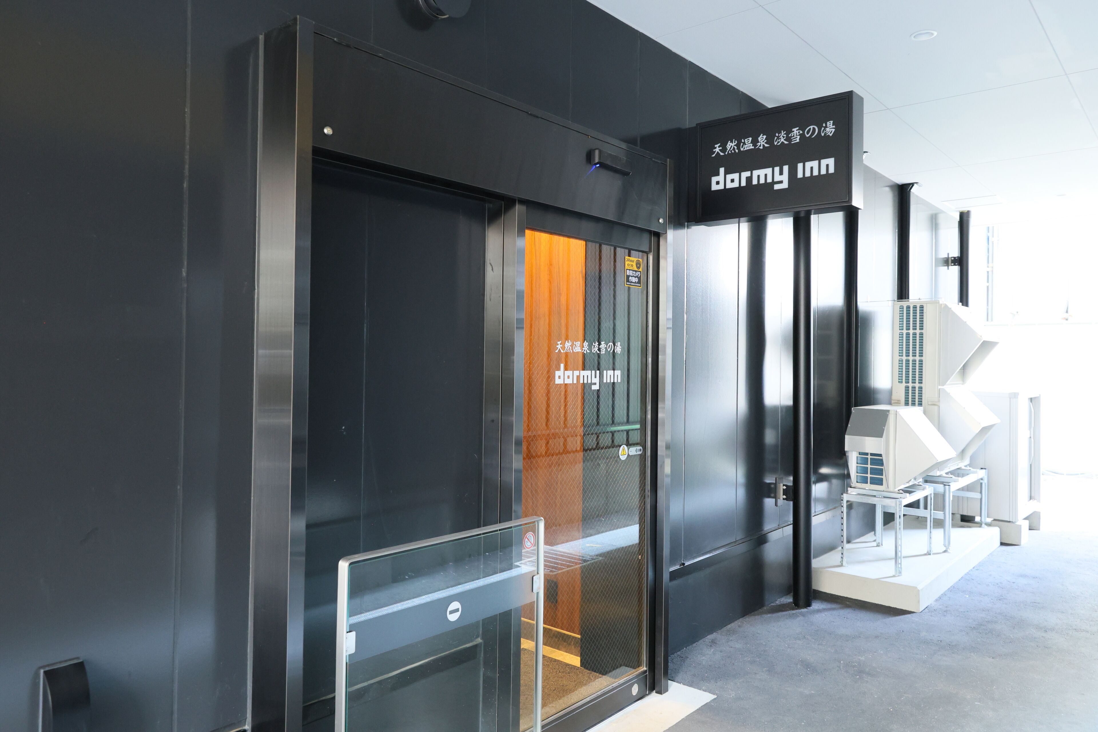 A modern building entrance with a 'dormy inn' sign, a glass door, and a small model house outside.
