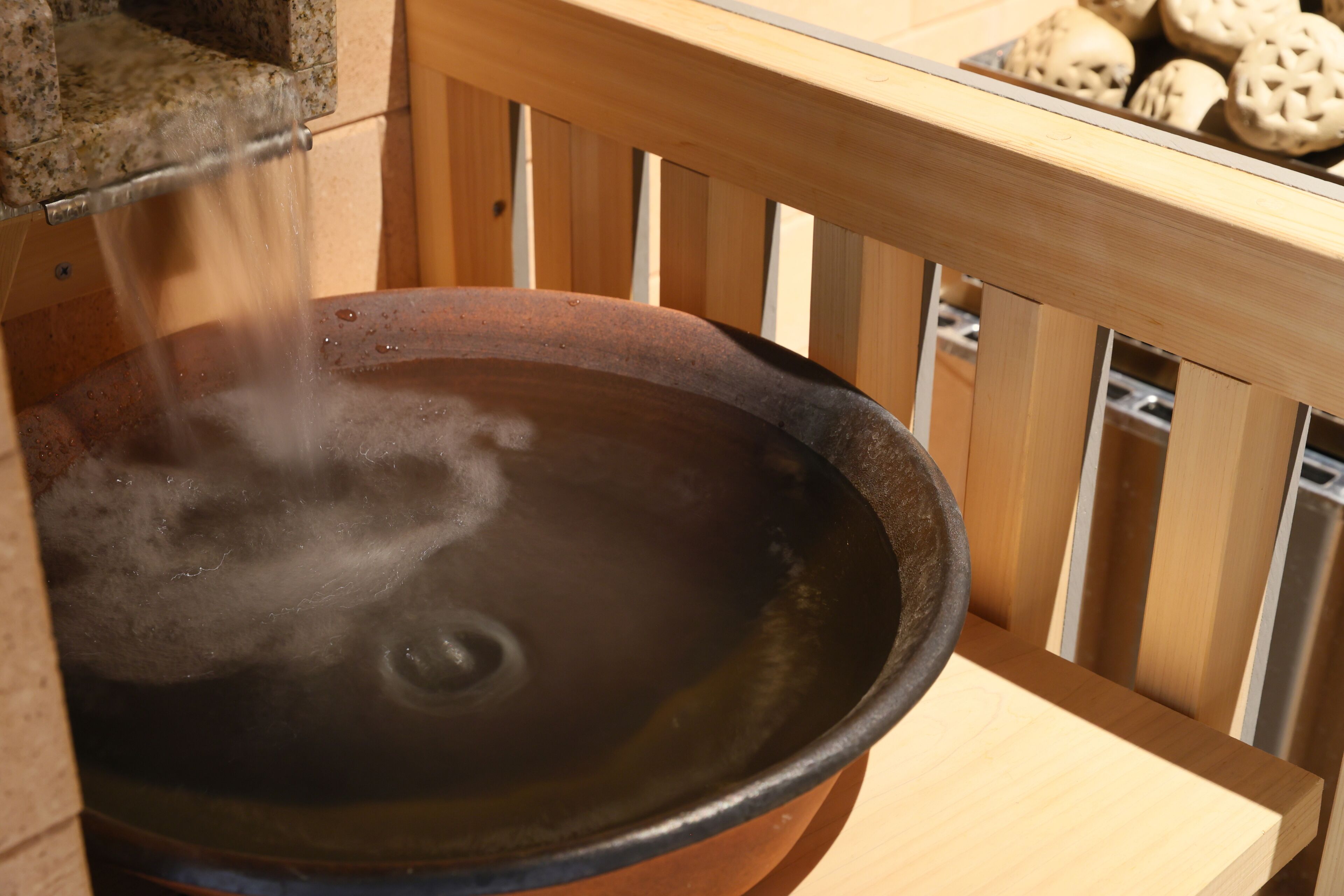 A hot spring bath with steam rising from the water, wooden railings, and a wooden table with baked goods in the background.