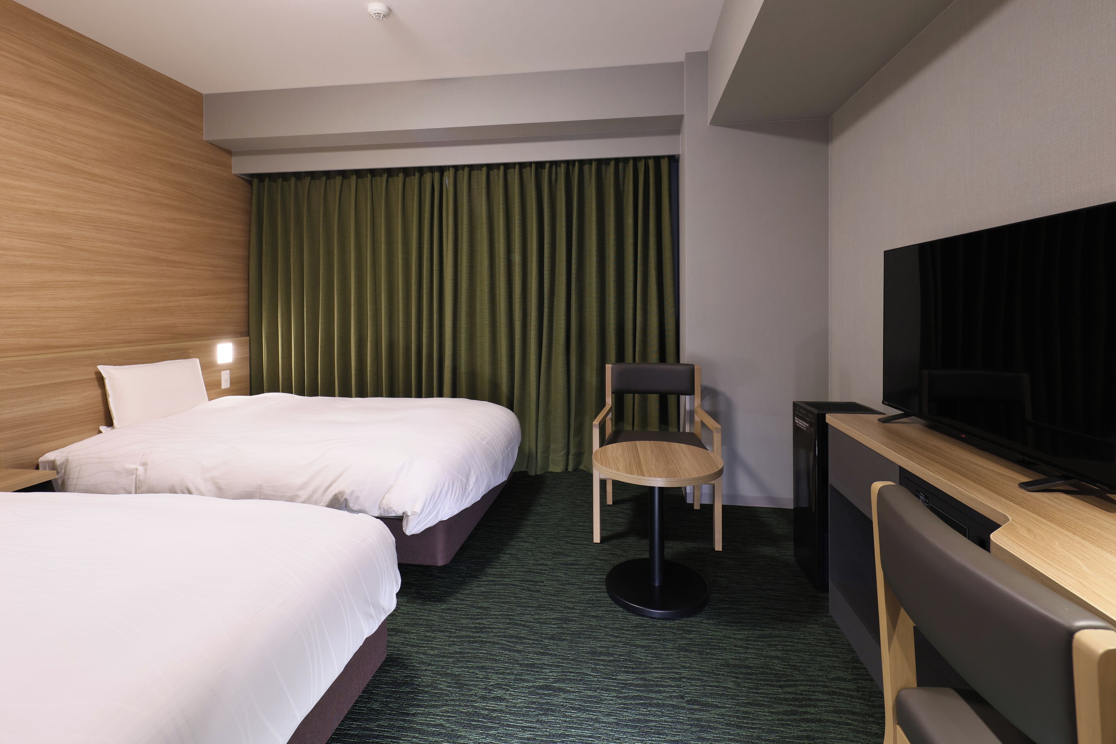 A hotel room with two beds, a desk with a TV, a chair, and a green curtain.