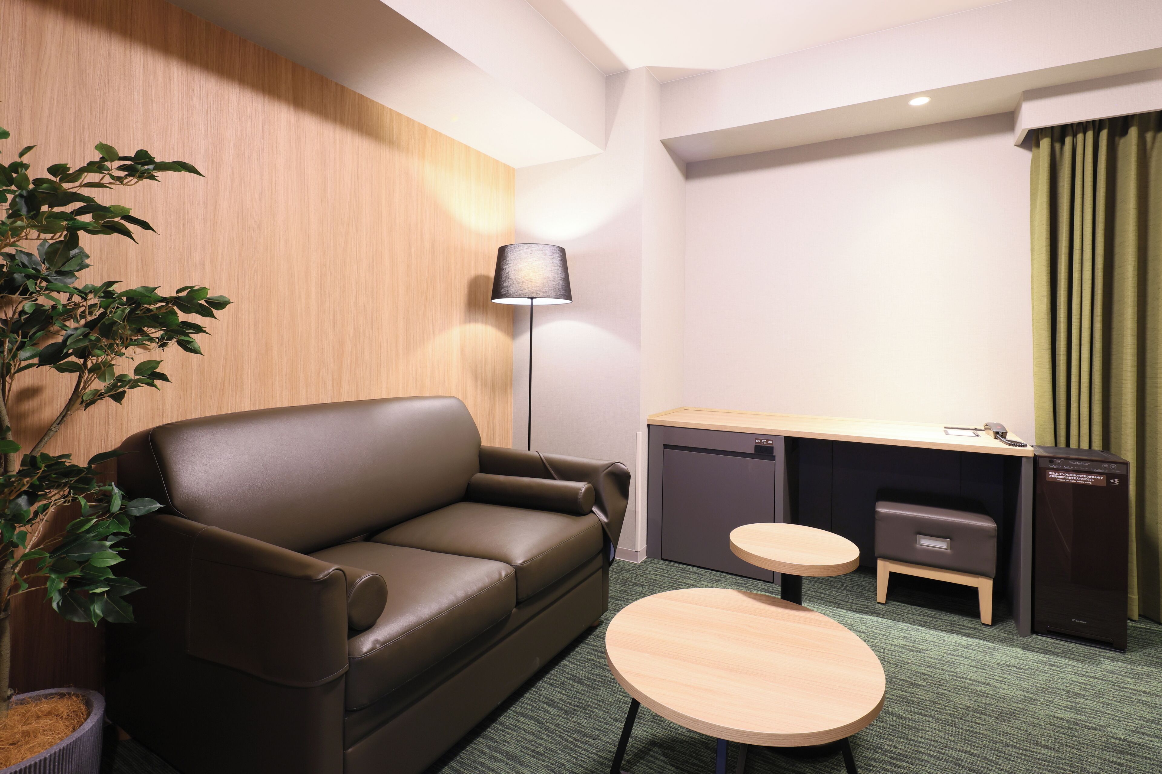 Une chambre d’hôtel moderne, dotée d’un canapé en cuir, d’une petite table ronde, d’un bureau avec un ordinateur, d’une lampe et d’une plante verte.