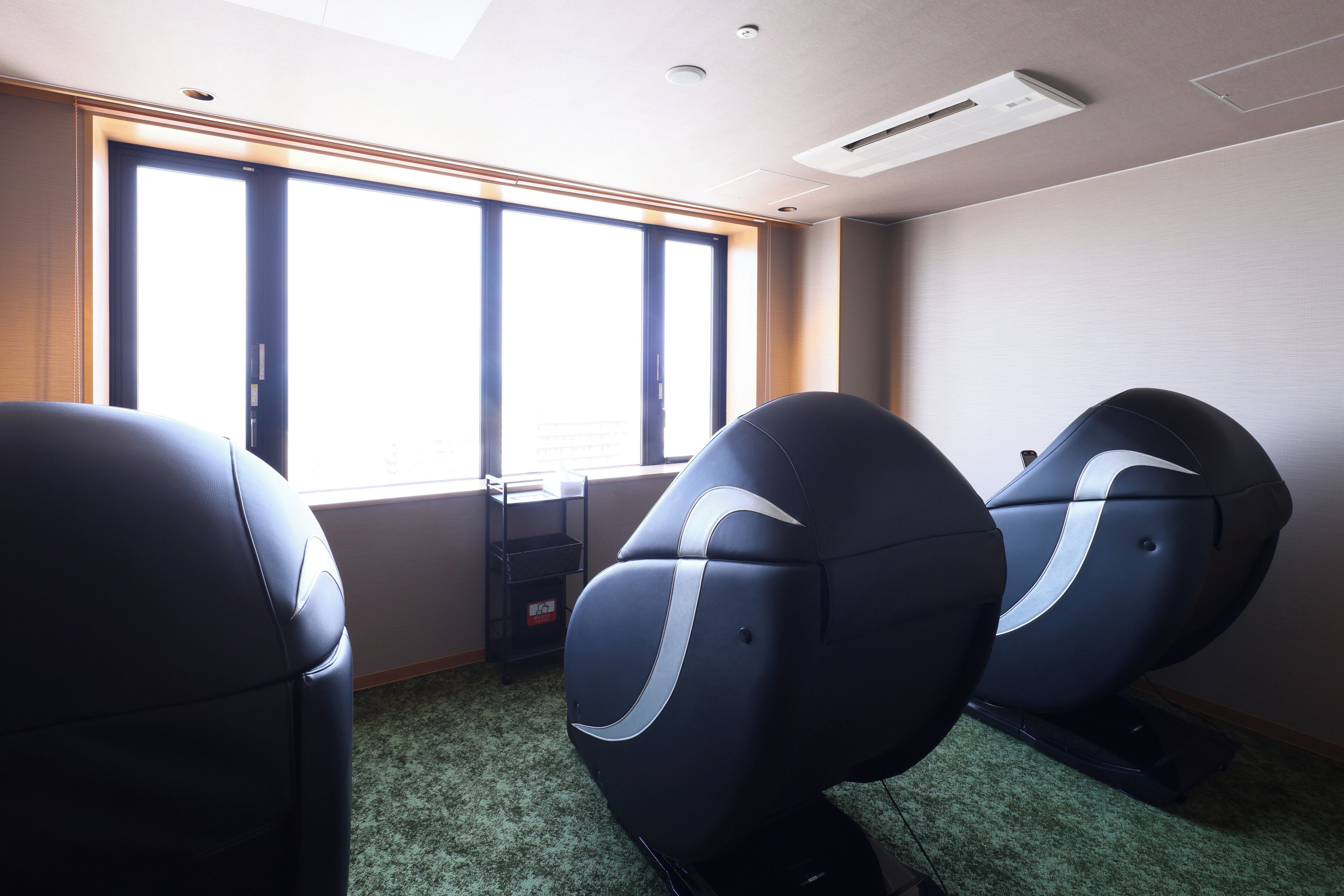A room with three massage chairs, a window with blinds, and a ceiling vent.