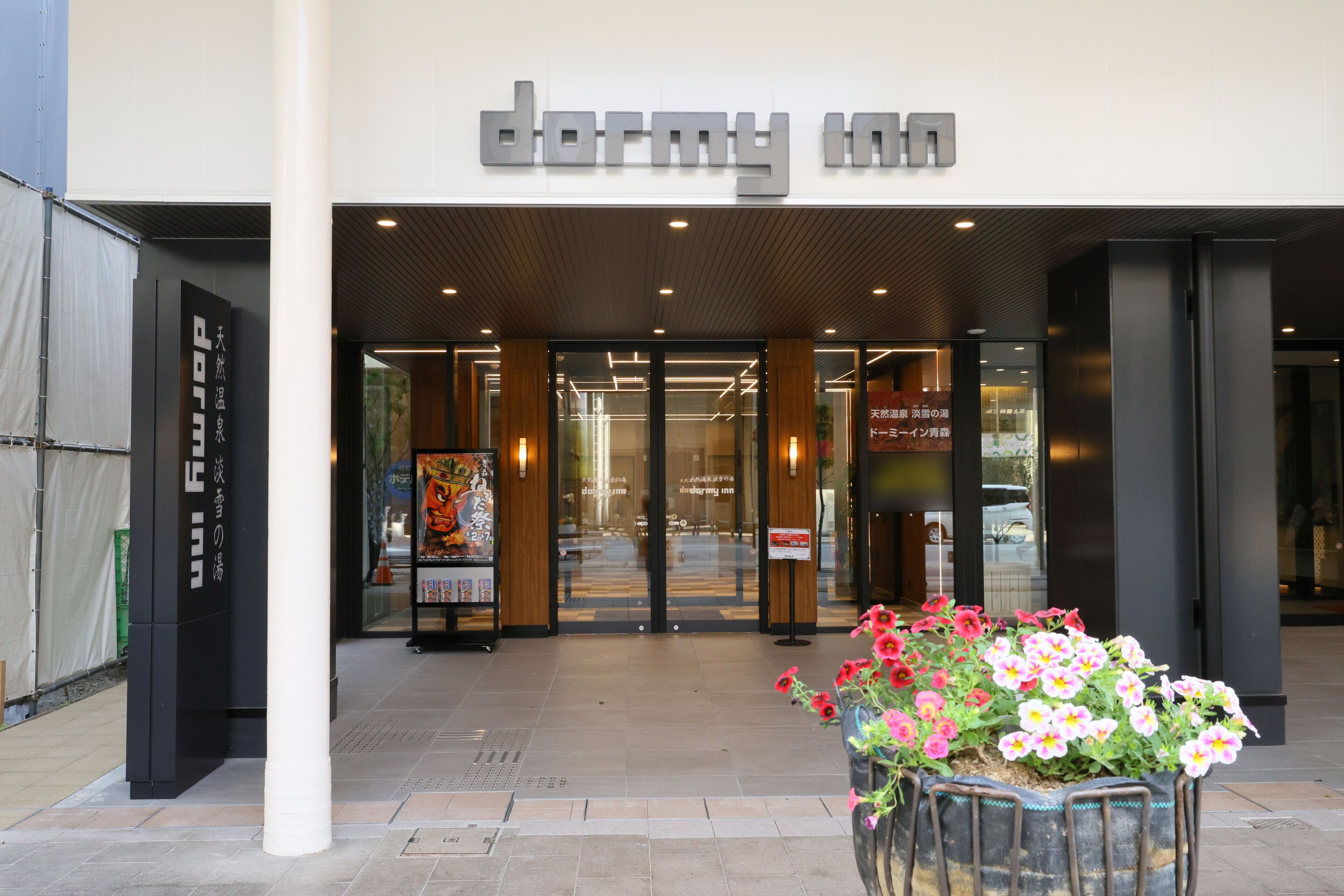 The entrance to a hotel with a modern facade, a signboard, and a flower planter in front.