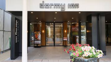 The entrance to a hotel with a modern facade, a signboard, and a flower planter in front.