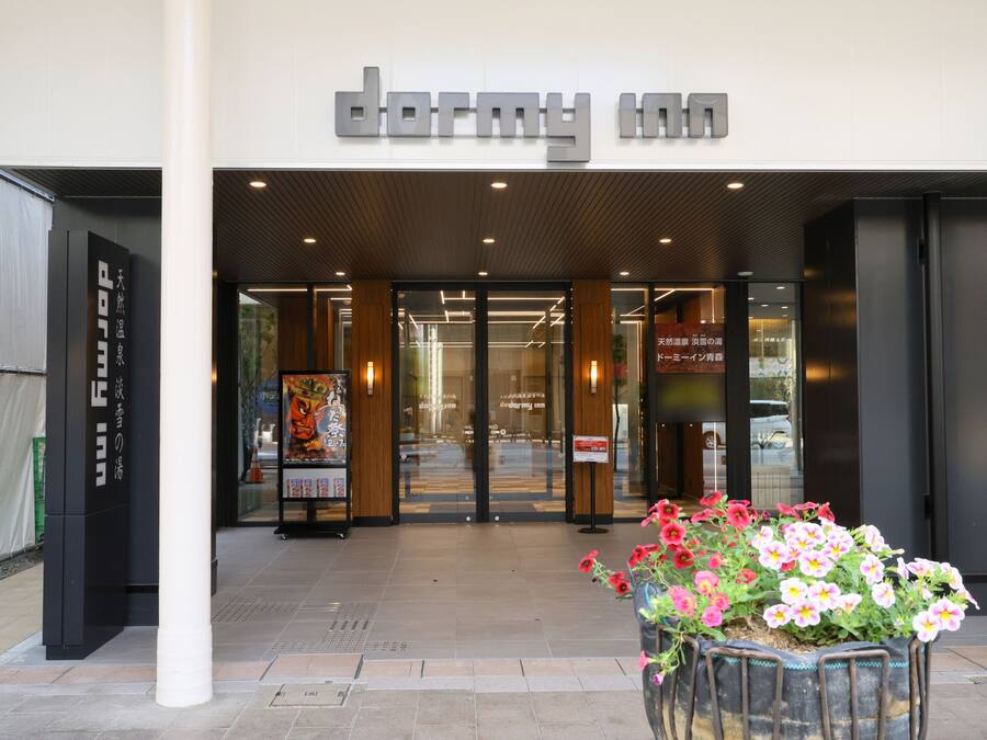 The entrance to a hotel with a modern facade, a signboard, and a flower planter in front.
