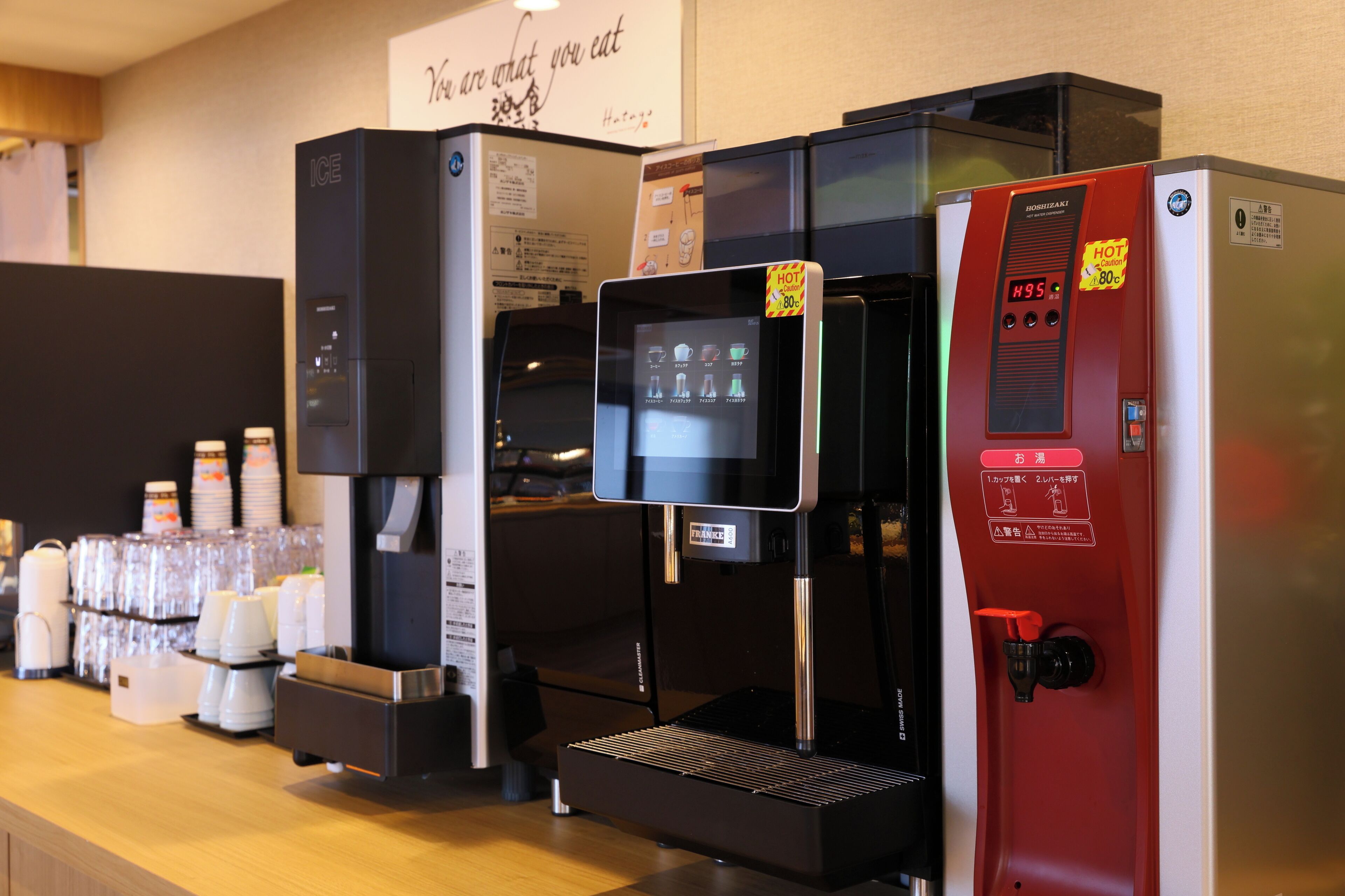 A coffee machine with a digital display and a red panel, next to a refrigerator and a stack of white cups.