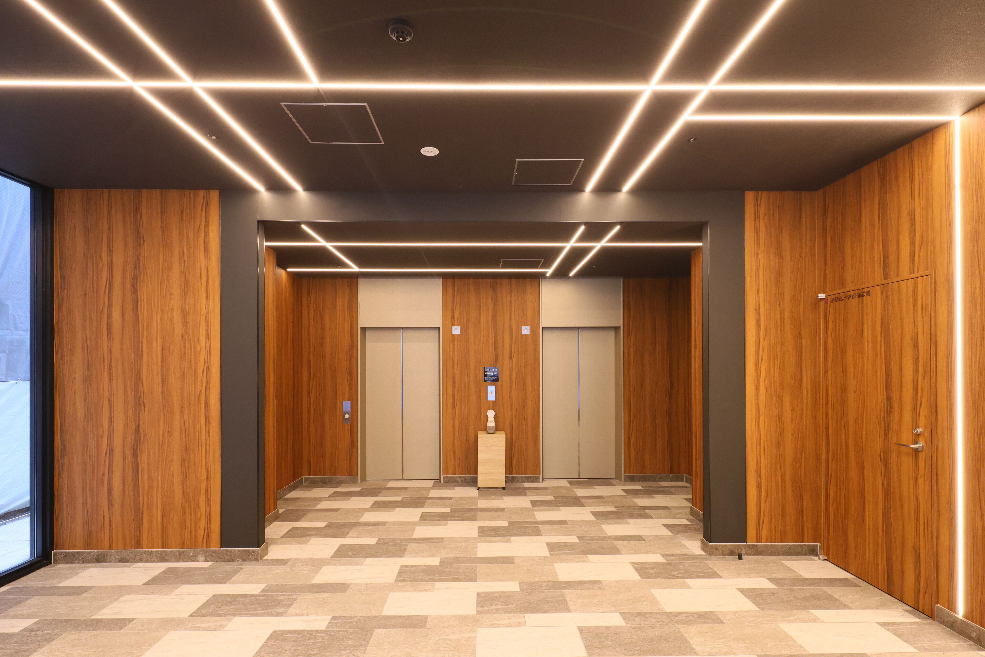 A modern elevator area with wooden paneling, a herringbone-patterned floor, and a central pillar with a plaque.