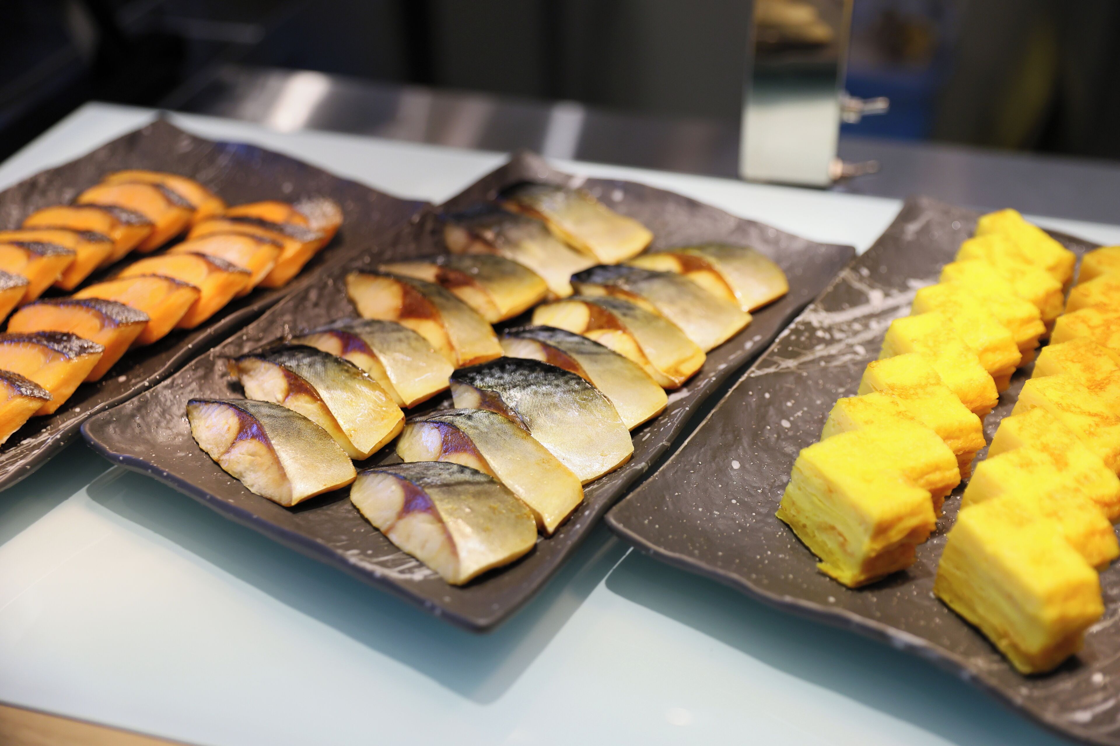 A buffet with various dishes including sliced fish, eggplant, and yellow cubes.