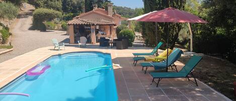 A swimming pool with lounge chairs and an umbrella, surrounded by a garden and a building with a tiled roof.