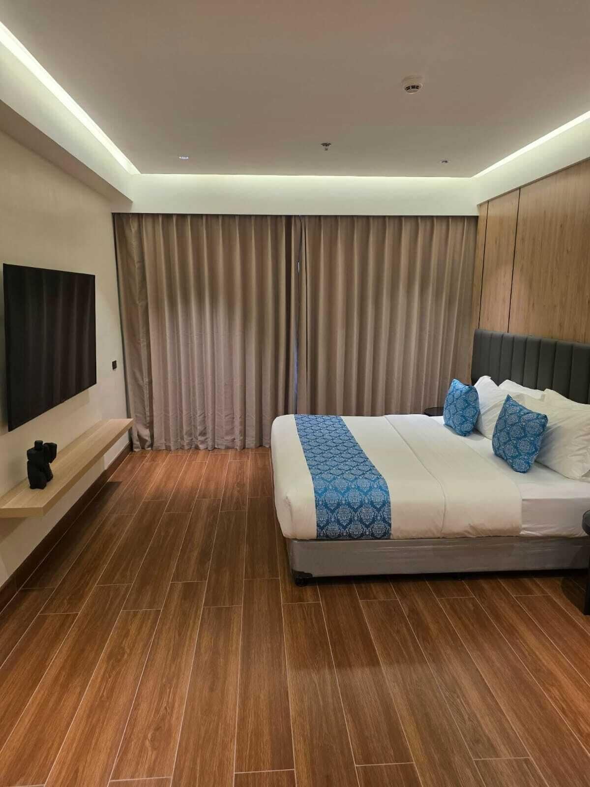 A modern hotel room with a large bed, a flat-screen TV mounted on the wall, and a wooden floor.