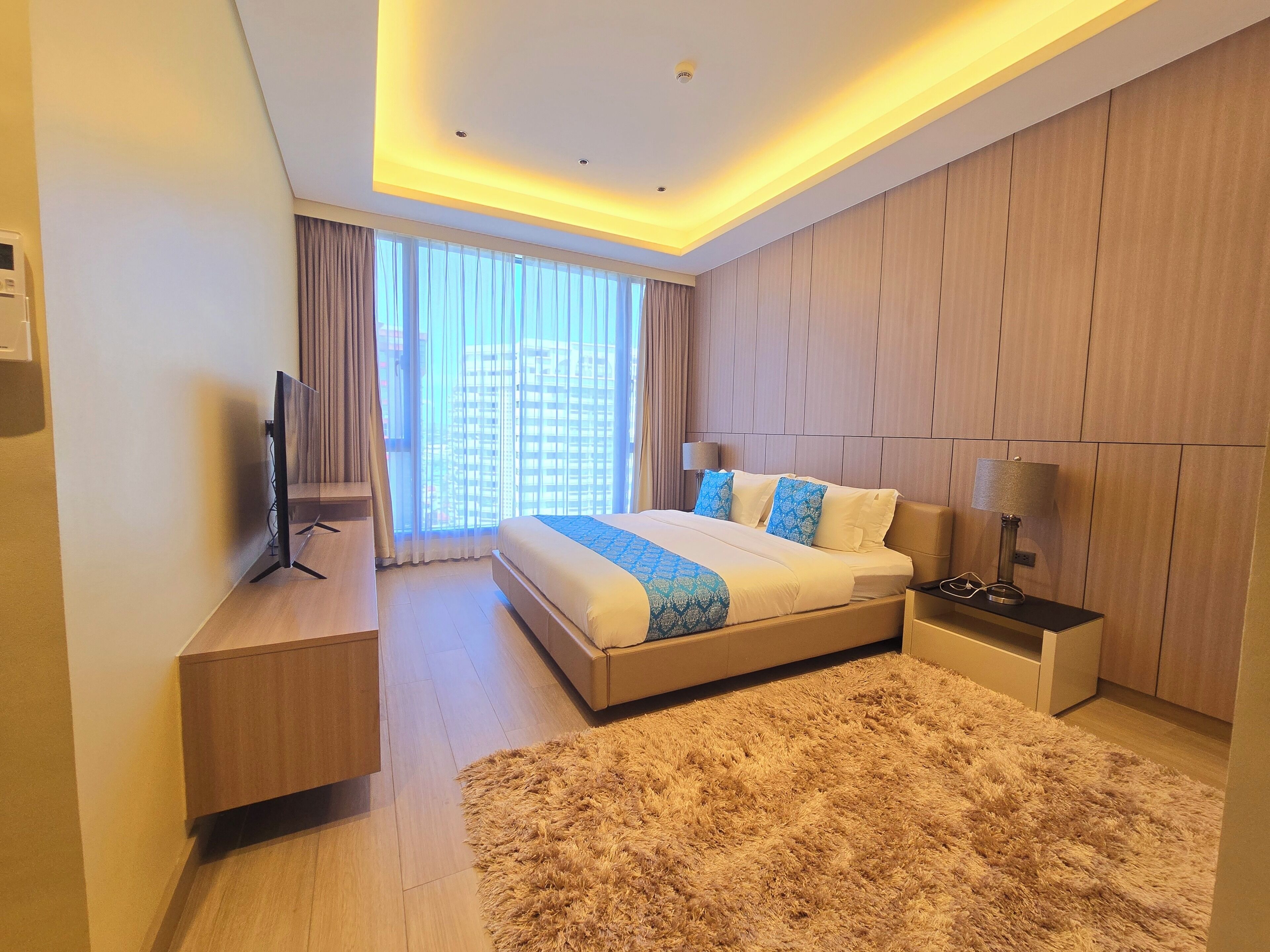 A modern hotel room with a large bed, a flat-screen TV, a bedside table, and a fluffy rug.