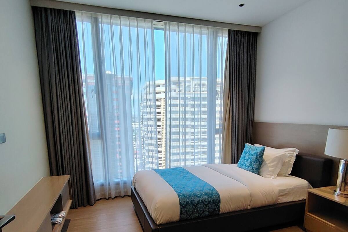 A modern hotel room with a large bed, a wooden nightstand, a desk, and a view of tall buildings through sheer curtains.