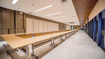 Una sala conferenze ampia e moderna con file di tavoli e sedie, un soffitto alto con illuminazione lineare e un pavimento pulito e lucido.