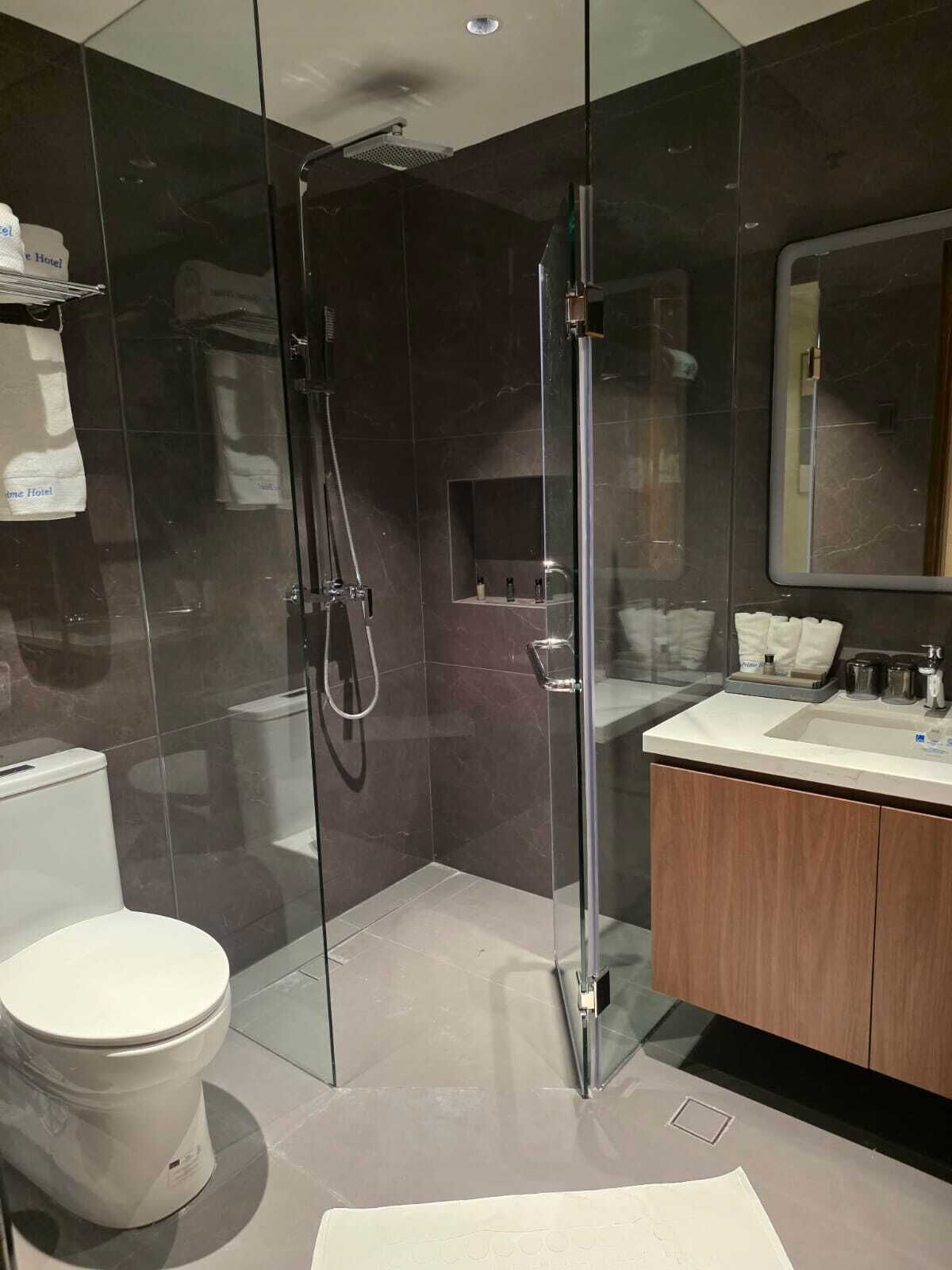 Un baño moderno con ducha envuelta en vidrio, inodoro, tocador con espejo y repisa con artículos de aseo.