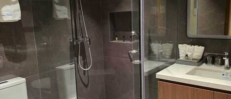 Un baño moderno con ducha envuelta en vidrio, inodoro, tocador con espejo y repisa con artículos de aseo.