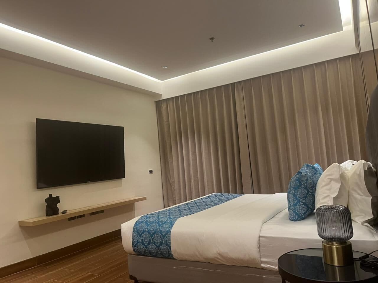 Ein modernes Hotelzimmer mit einem wandmontierten Flachbildfernseher, einem Bett mit gemustertem Überwurf, einem Nachttisch mit Lampe und einem Fenster mit Vorhängen.