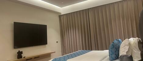 Una habitación de hotel moderna con una televisión de pantalla plana montada en la pared, una cama con colcha estampada, una mesita de noche con lámpara y una ventana con cortinas.
