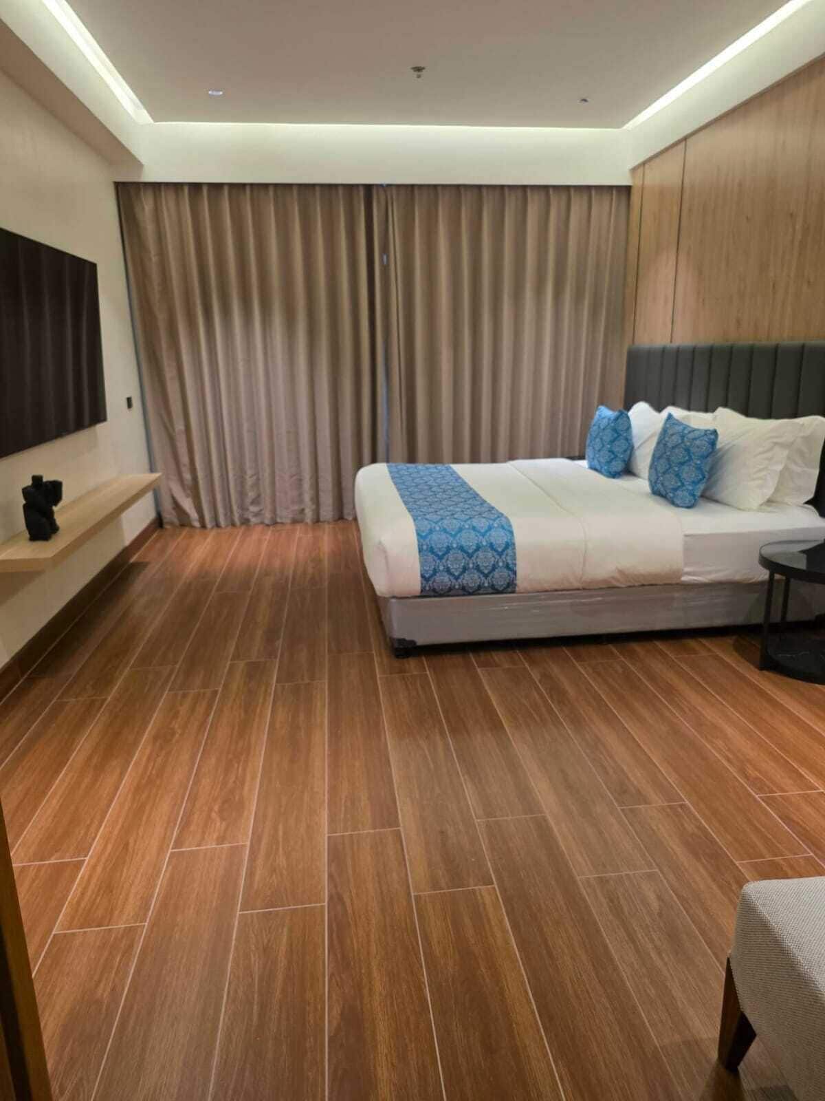 Ein modernes Hotelzimmer mit einem großen Bett, Holzboden, einem Flachbildfernseher und einem großen Fenster mit Vorhängen.