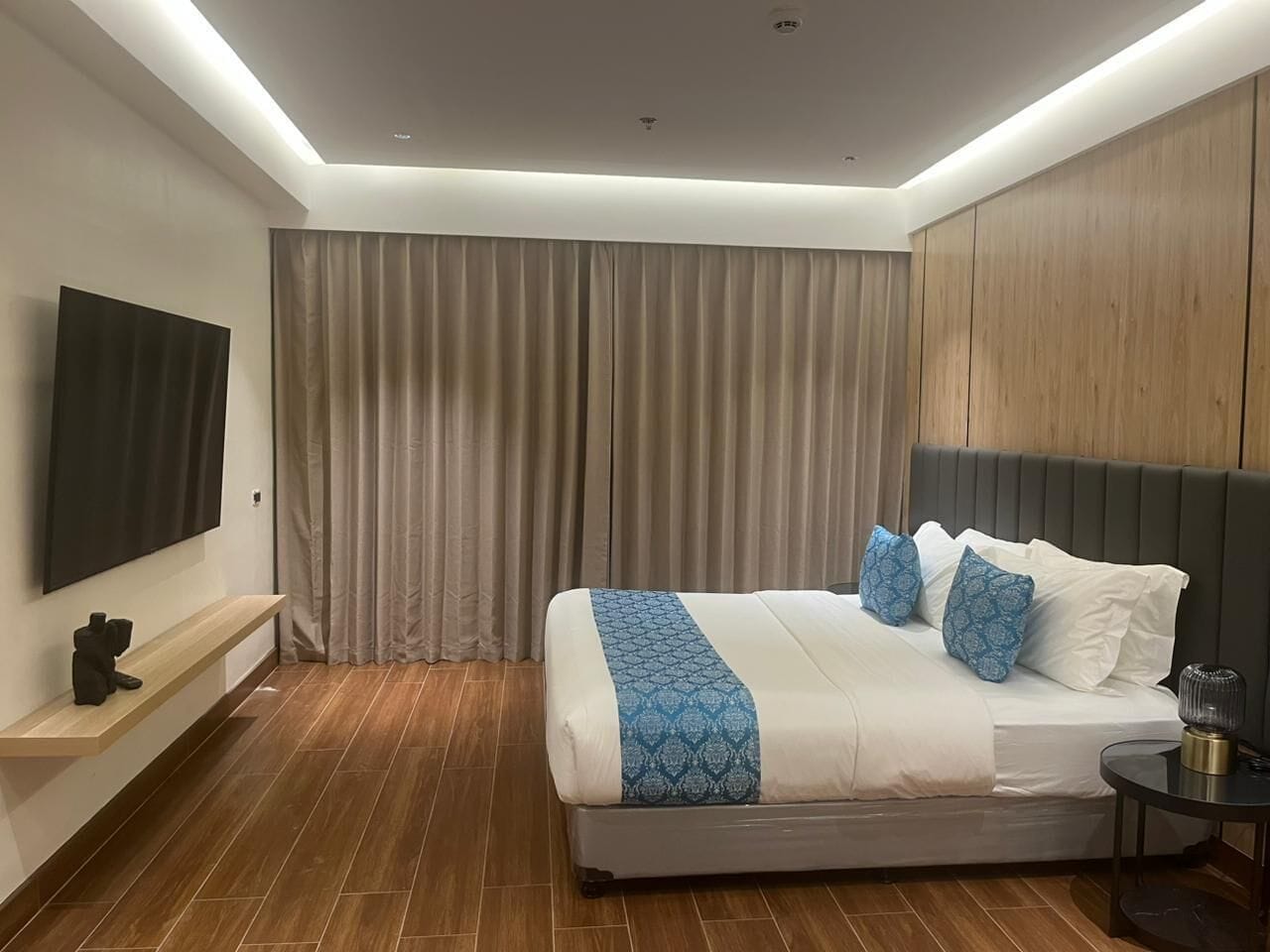 A modern hotel room with a large bed, a flat-screen TV mounted on the wall, a wooden headboard, and a wooden floor.