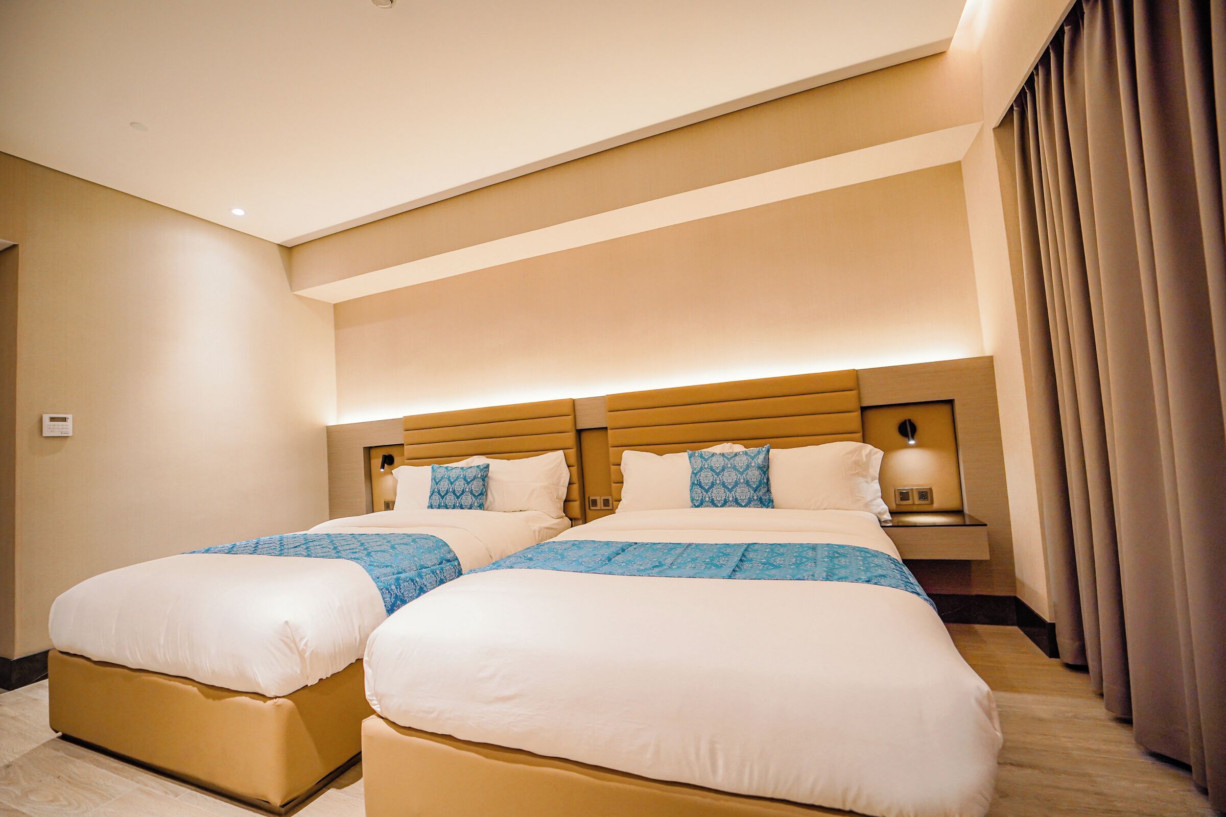 A hotel room with two beds, a wooden headboard, a nightstand with a lamp, and a window with curtains.