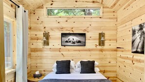 Een slaapkamer in een houten chalet, met een bed, twee wandlampen, een ingelijste foto en een raam met gordijnen.