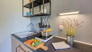 Una cocina con un lavamanos, una tabla de cortar con verduras, una olla sobre una placa de inducción y una planta en maceta.