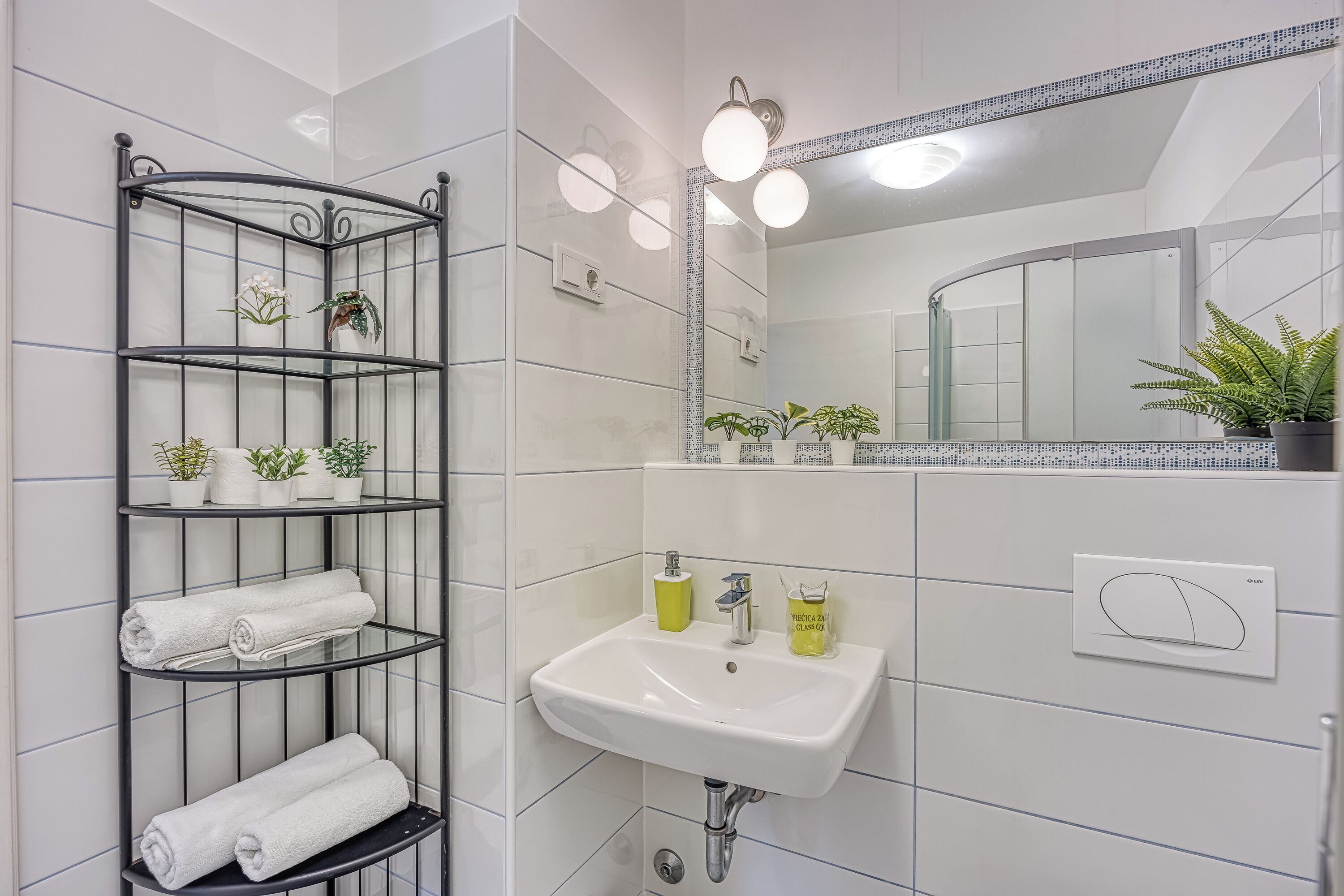 A bathroom with a white tiled wall, a black metal shelf with towels and plants, a white sink with a mirror above it, and a light fixture.