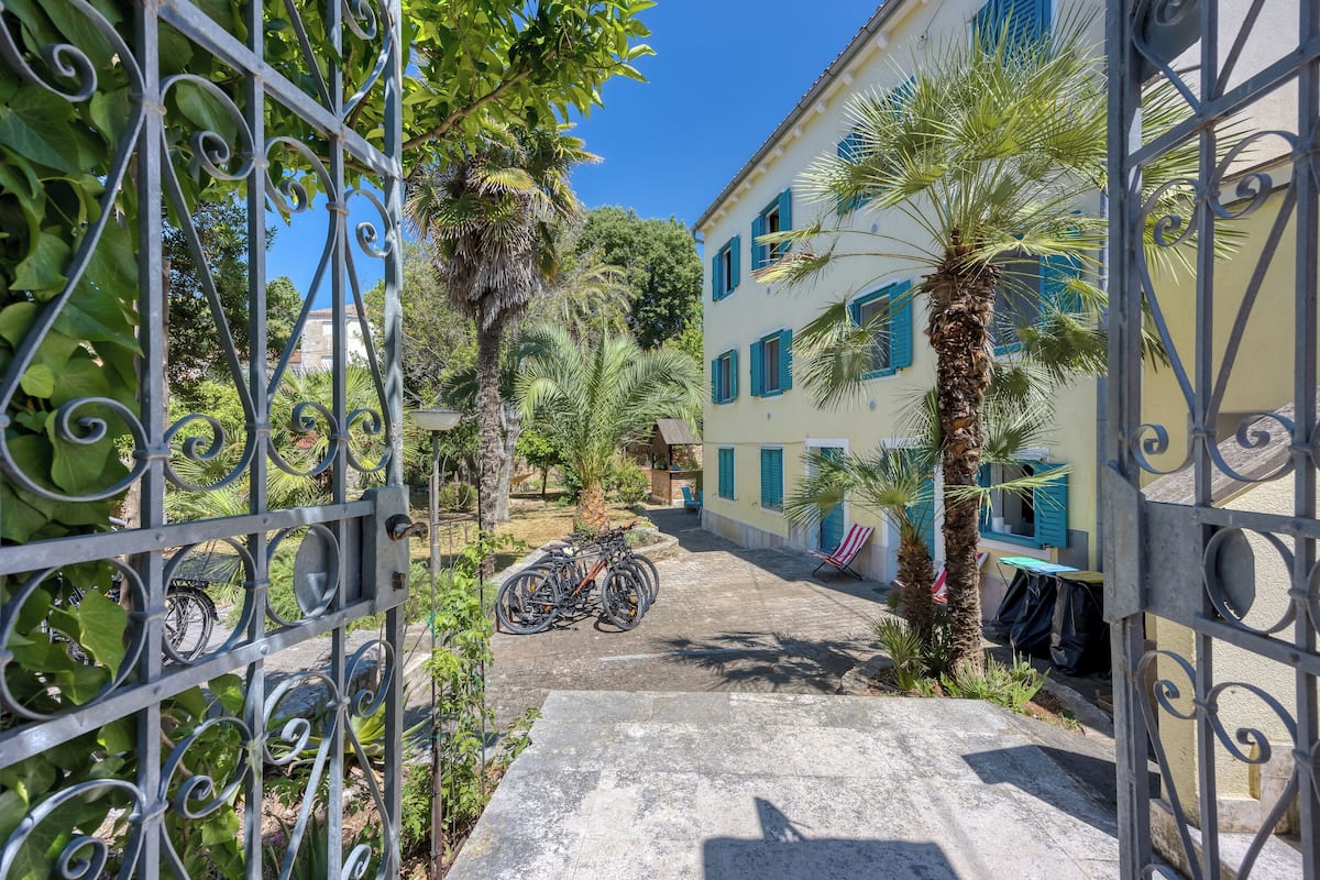 A courtyard with a paved path leading to a building with blue shutters, surrounded by greenery and a metal gate.