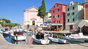 Un puerto deportivo con varias embarcaciones amarradas, un edificio rosado de arquitectura clásica, y edificios coloridos al fondo.