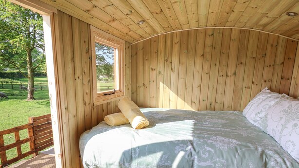 A wooden cabin room with a bed, a window, and a view of a grassy area with trees. - The Oak Hut (Whitchurch)