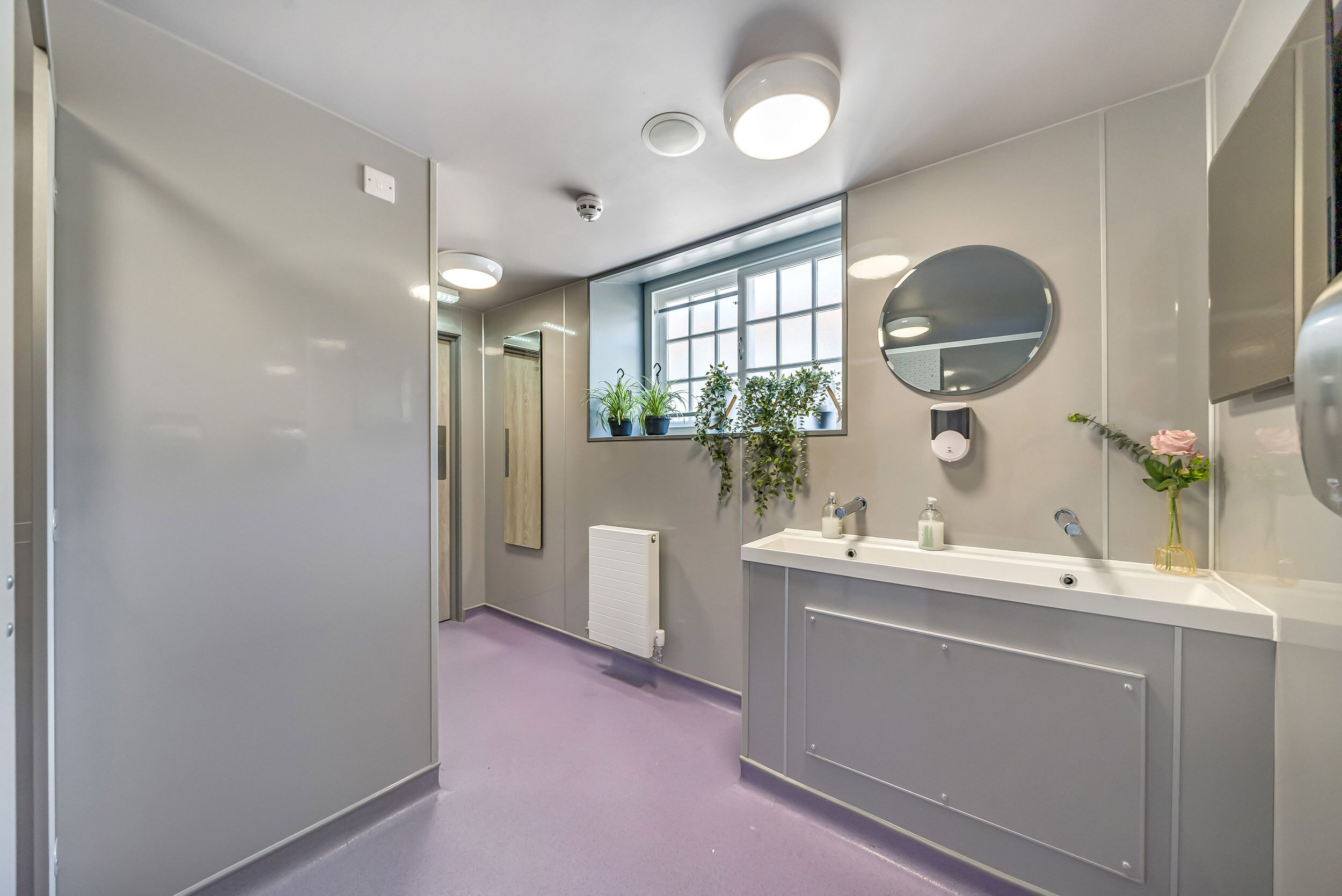 A modern bathroom with a large mirror, two sinks, and a window with plants.