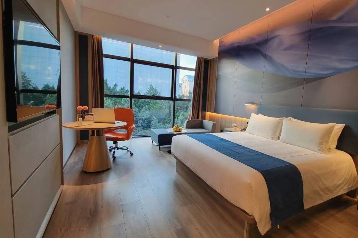 A modern hotel room with a large bed, a desk with a chair, a small table with a lamp, and a view of the outdoors through large windows.