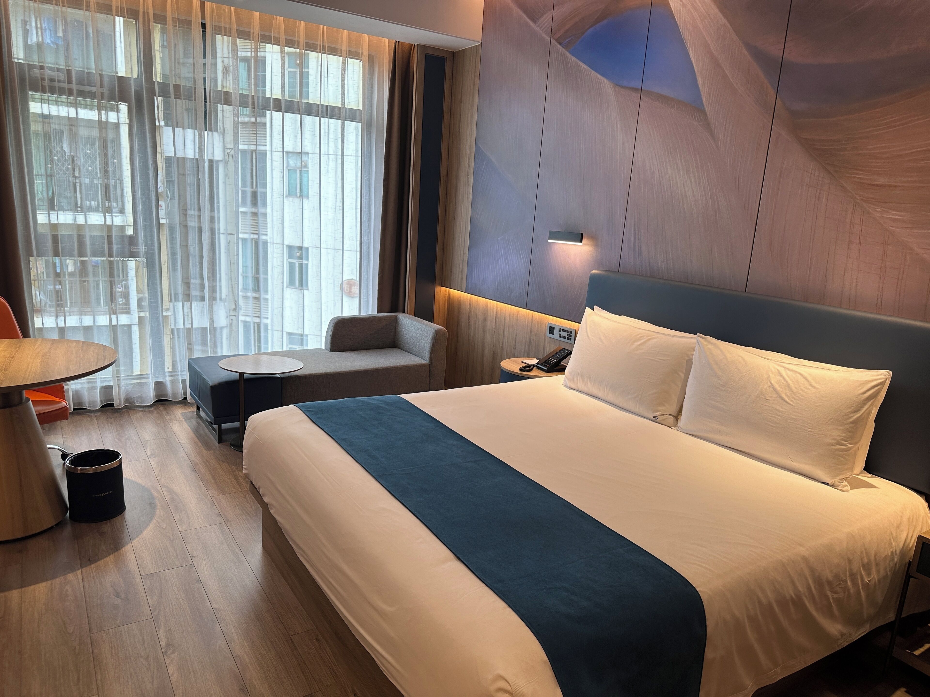 Habitación de hotel con una cama grande, una mesa redonda pequeña, un sofá y una ventana con cortinas traslúcidas.